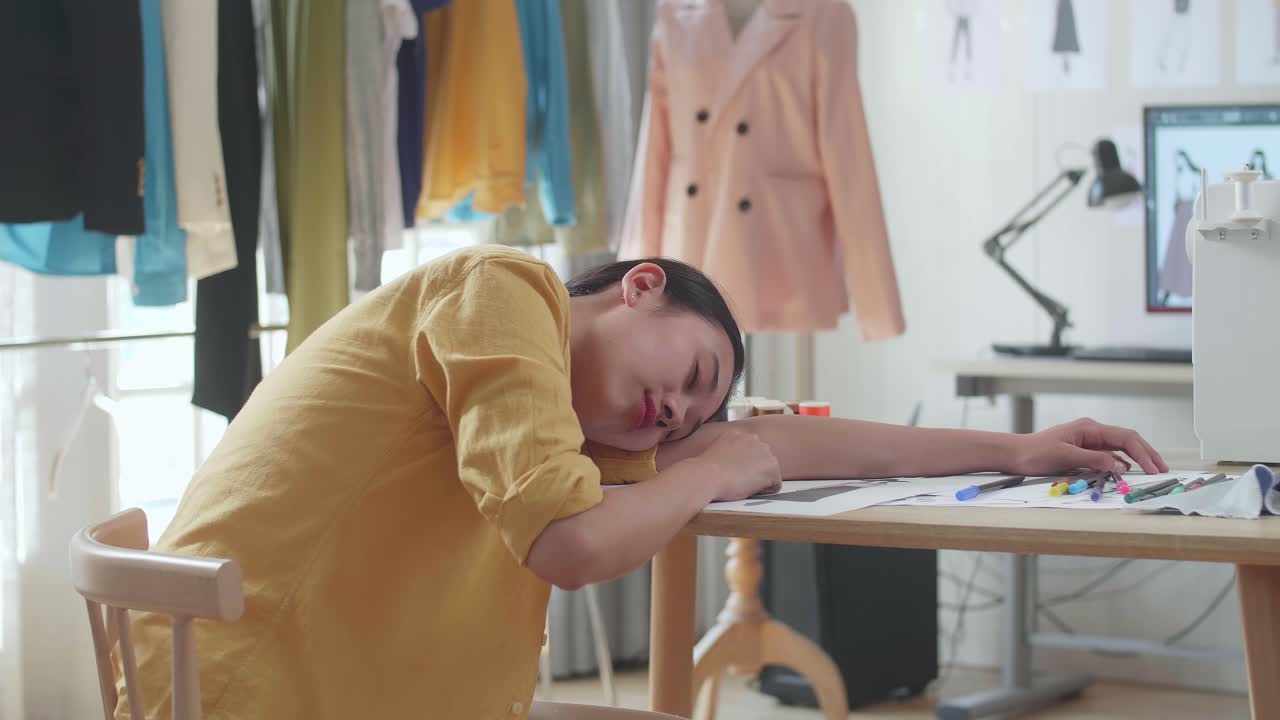 아시아 여성 디자이너가 바느질 기계와 함께 스튜디오에서 옷을 디자인하면서 잠을 자고 있습니다.