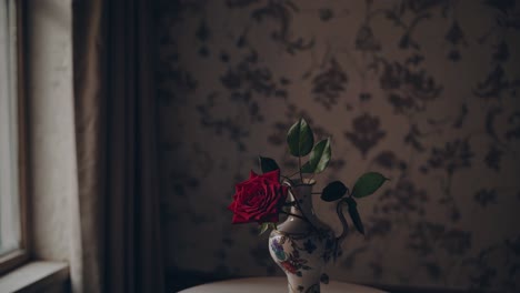 red rose with green leaves in vintage vase resting on a small white table near window with curtains and wallpaper in the background, creating a nostalgic and moody atmosphere