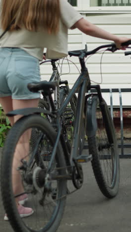 back view of young woman in pink sneakers, jean shorts, and black handbag moving bicycle toward bicycle stand, setting bike among other parked bicycles with white building backdrop