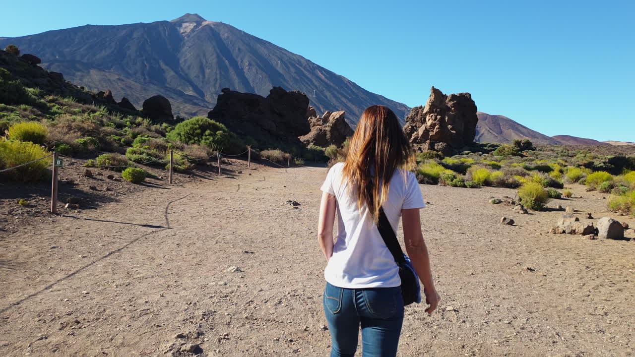 Solitary female traveler walking leisurely along volcanic trail in Teide national park, capturing serene landscape with rocky terrain and blue sky during summer vacation