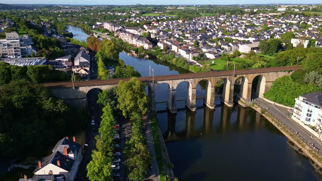 Drone shot over Laval, France—arched stone viaduct crosses Mayenne River, connecting historic city center, green riverbanks, and white-roofed homes under a clear sky