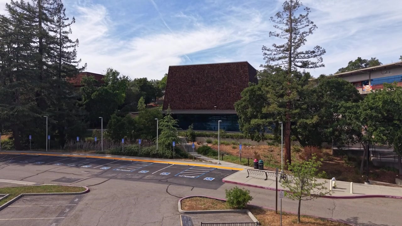 Toma aérea con dron ascendiendo sobre un estacionamiento casi vacío, revelando la arquitectura moderna del campus de Foothill College, enclavado entre árboles verdes en Los Altos Hills, California, en un día soleado