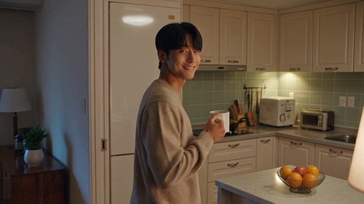 Uomo adulto con un maglione leggero che cammina verso una cucina aperta per prendere da bere, tenendo una tazza bianca.