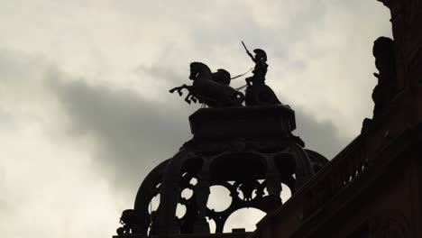 Poderosa-Escena-Cinematográfica-De-Un-Guerrero-Romano-Montando-Un-Carro-Con-Dos-Caballos-Con-El-Brazo-Levantado-Sosteniendo-Una-Espada-En-El-Cielo-Escultura-Encima-De-Una-Silueta-De-Techo-Nubes-Dramáticas-Moviéndose-A-Alta-Velocidad-De-Fondo