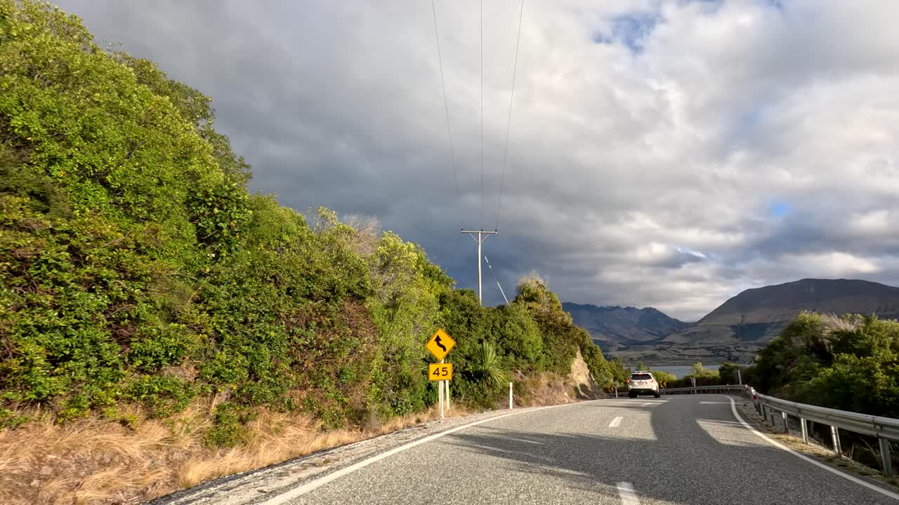 Car travels scenic mountain road with lush greenery, dramatic clouds, and afternoon sunlight in Queenstown