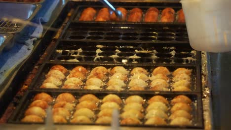 a street food vendor prepares takoyaki at the local night market, adding ingredients to the fillings of these delicious round octopus balls, close up shot.