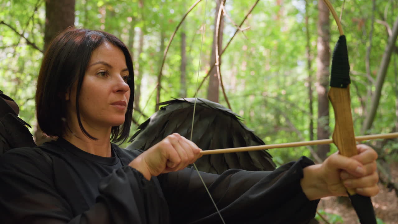 Close up side view of female archer with black wings holding bow and arrow in forest, sunlight filtering through leaves, showing calm focus, confidence, and strength in mystical natural woodland