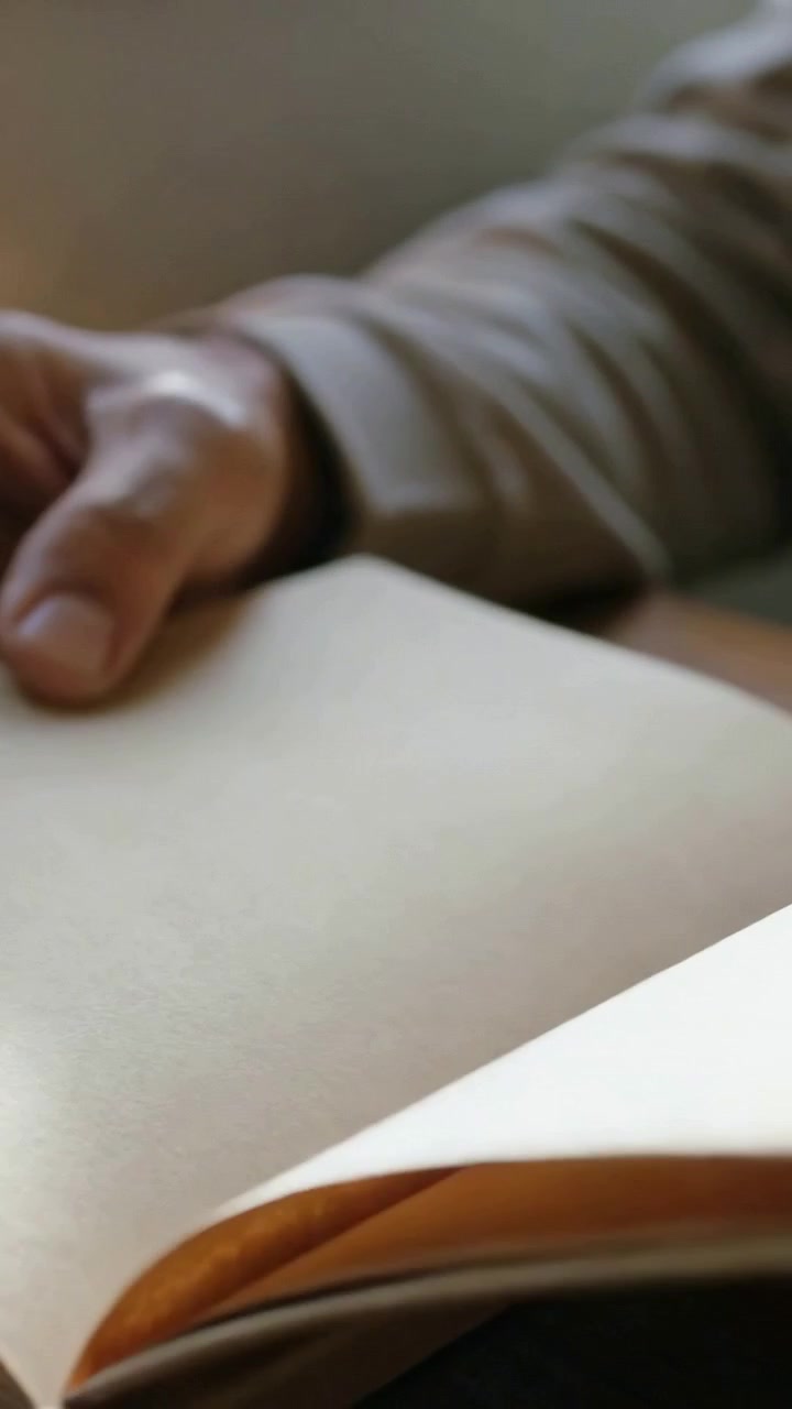 Video verticale: Sollevando il bordo, uomo con manica beige che sfoglia un quaderno kraft al tavolo, mentre prende appunti