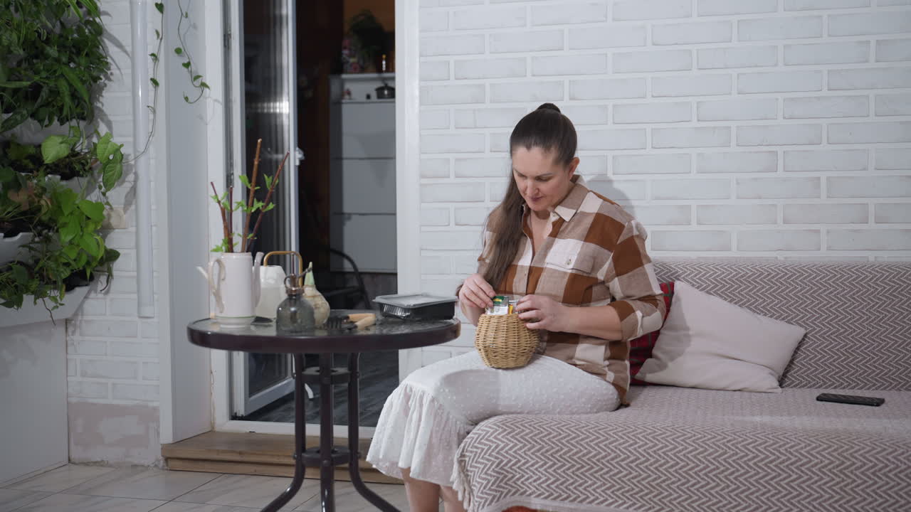 Urban gardener carrying wicker basket filled with seed packets, walking toward sofa in cozy indoor garden space, surrounded by leafy plants on white brick wall backdrop, wearing checkered shirt