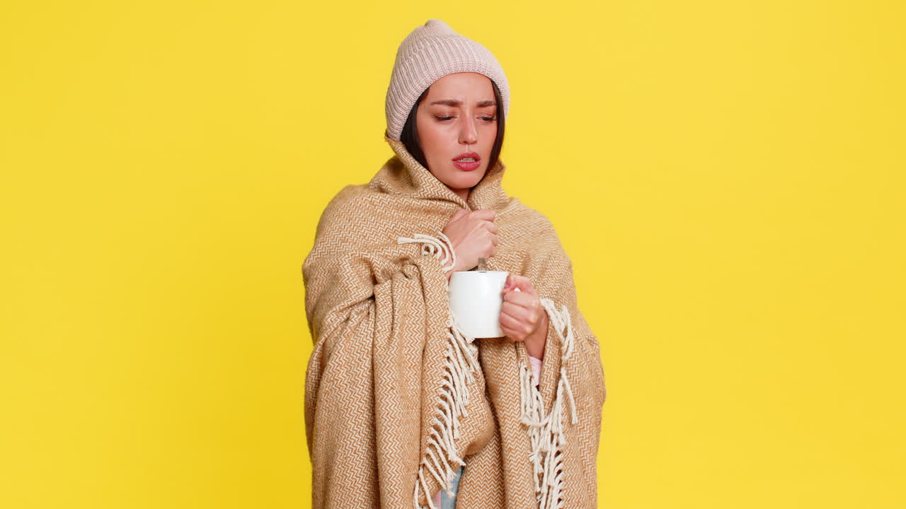 Woman wrapped in blanket wearing hat adding honey to tea slowly drinking caring for sore throat