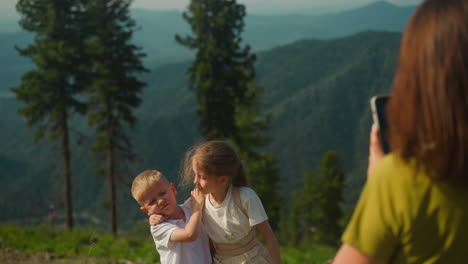 mom takes photos of kids with rugged mountains on background. family spends time together surrounded by pristine nature. good relations of parents and children