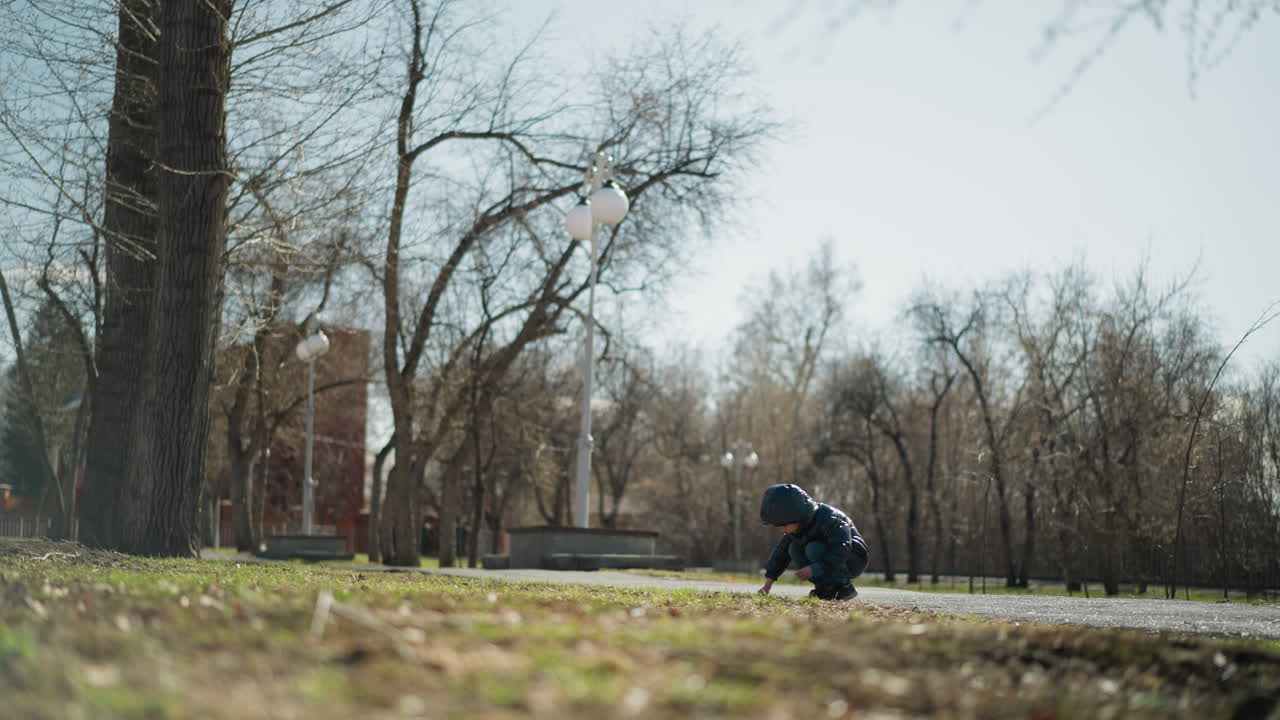 한 어린 소년이 손에 지이를 들고 을 파는 것을 볼 수 있습니다. 그는 검은 가죽 재, 헤드 워머, 그리고 청바지를 따뜻하게 입고 있습니다. 나무와 거리 램프가 주변에 있습니다.