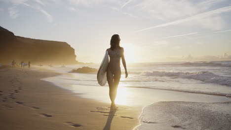 woman walking on the beach with surfboard at sunset
