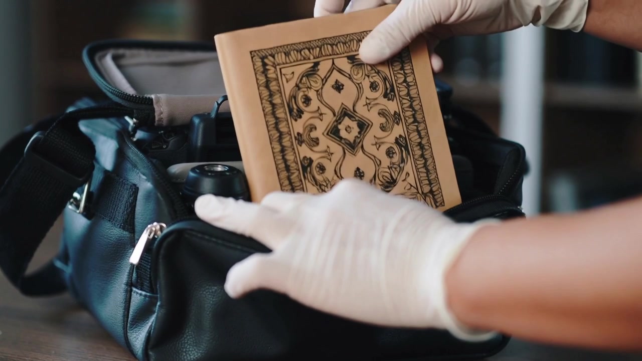 Entering from right on table, hands wearing gloves lowering tan book into camera bag for safety