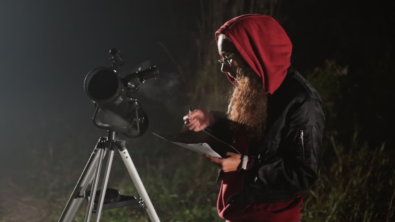 Hvid mand med skæg justerer teleskop, nærbillede i mørkt felt, tjekker tablet og fokus, rød hættejakke, stativmonteret teleskop, tåget atmosfære, metodisk kalibrering, amatørforsker observerer