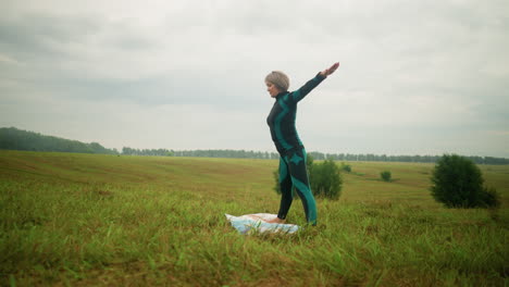 vista lateral de una mujer en traje negro verde de pie en una alfombra de yoga practicando la postura triangular hacia adelante, brazos extendidos, en un campo abierto de hierba bajo un cielo nublado con árboles y vegetación en la distancia