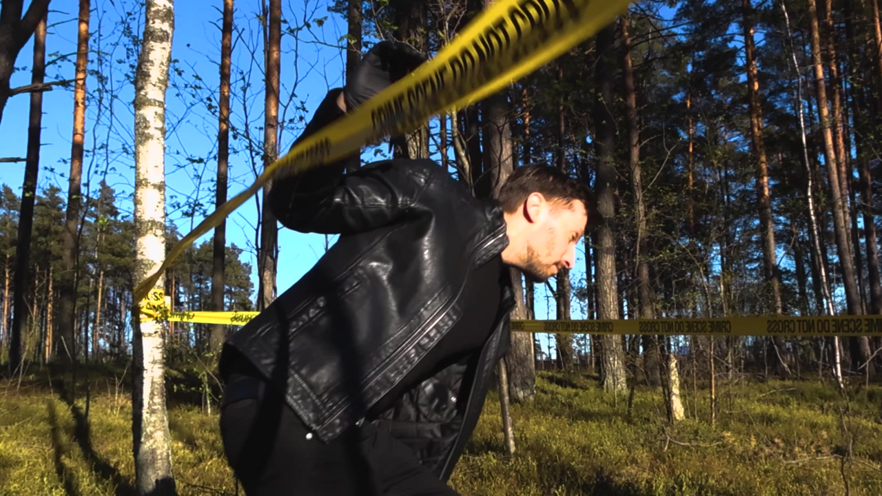 Sivil politibetjent eller detektiv i en svart skinnjakke som forsiktig krysser under det gule sperrebåndet på et åsted i en skog.