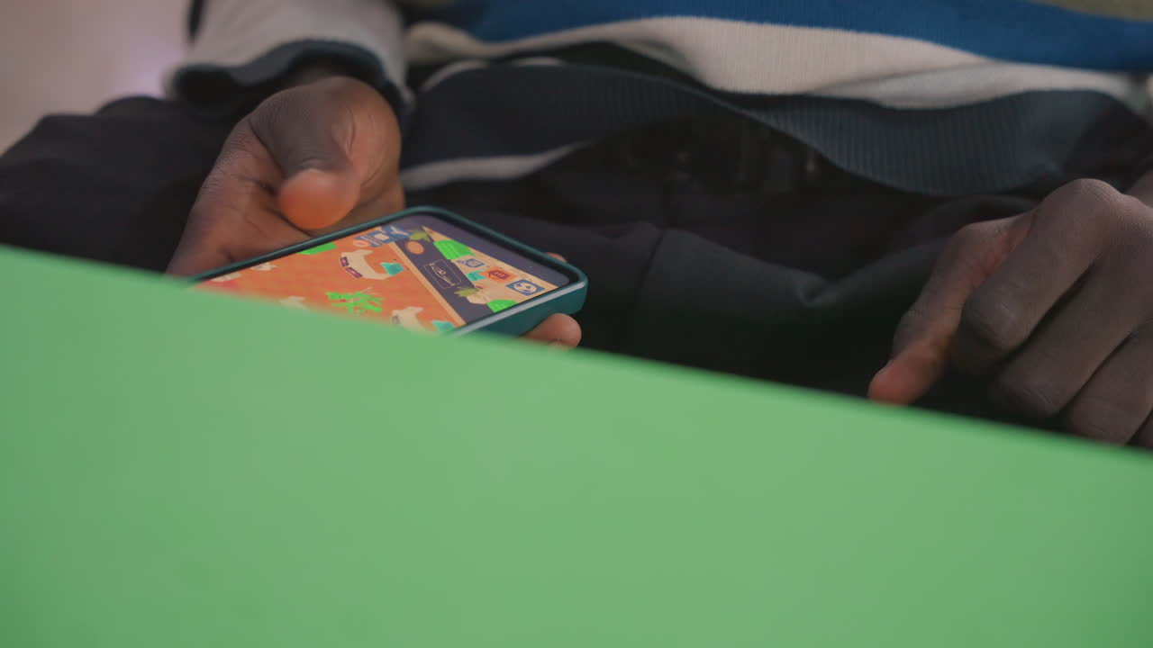 Mani scure che tengono il telefono, tavolo verde ritrae una persona di colore che messaggia durante un appuntamento romantico, primo piano del pollice sullo schermo colorato, custodia fantasia e polpastrelli, atmosfera accogliente e a bassa luce evoca attesa