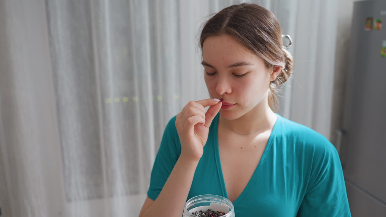 Young Woman Holding Supplement Jar, Contemplative Morning Scene With Teal Robe, Closeup Hands Picking Vitamin Capsule From Container, Soft Daylight Through Curtains, Calm Domestic Routine And Private