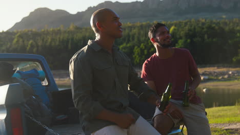 two male friends with backpacks in pick up truck on road trip by lake drinking beer