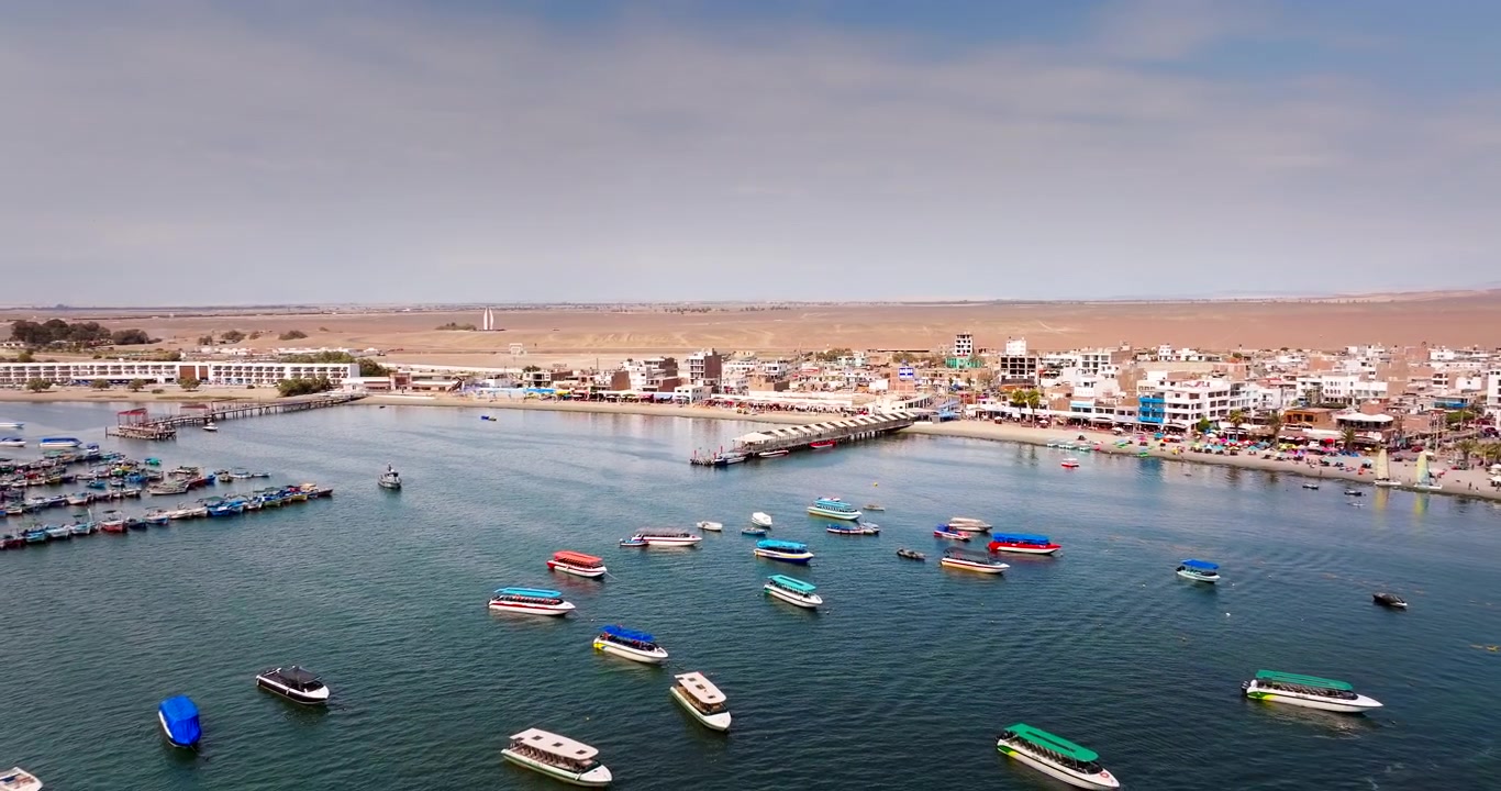 Numerose barche colorate ancorate in acque oceaniche calme vicino a un molo e a una città desertica a Paracas, Perù. Ripresa aerea all'indietro, spazio per il testo