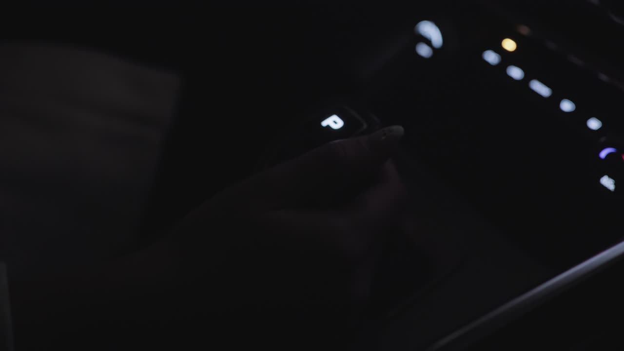 Close up of driver hand gripping illuminated gear selector in park mode inside moving car at night, with soft golden light reflecting on hand and dashboard as vehicle begins motion
