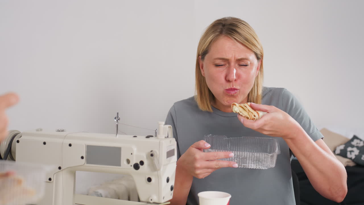 Needle women seated beside sewing machines in bright tailoring workspace eating pizza, one woman showing visible satisfaction as she takes a bite, enjoying casual break time surrounded by fabrics
