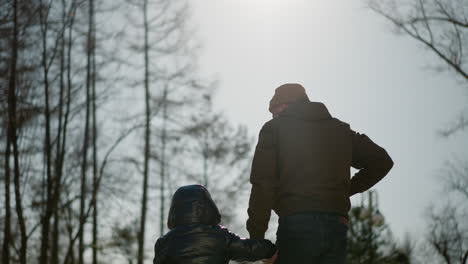 a father and son walk hand in hand outdoors, with the father putting his right hand into his jacket pocket as they walk, with trees and a bright sky in view