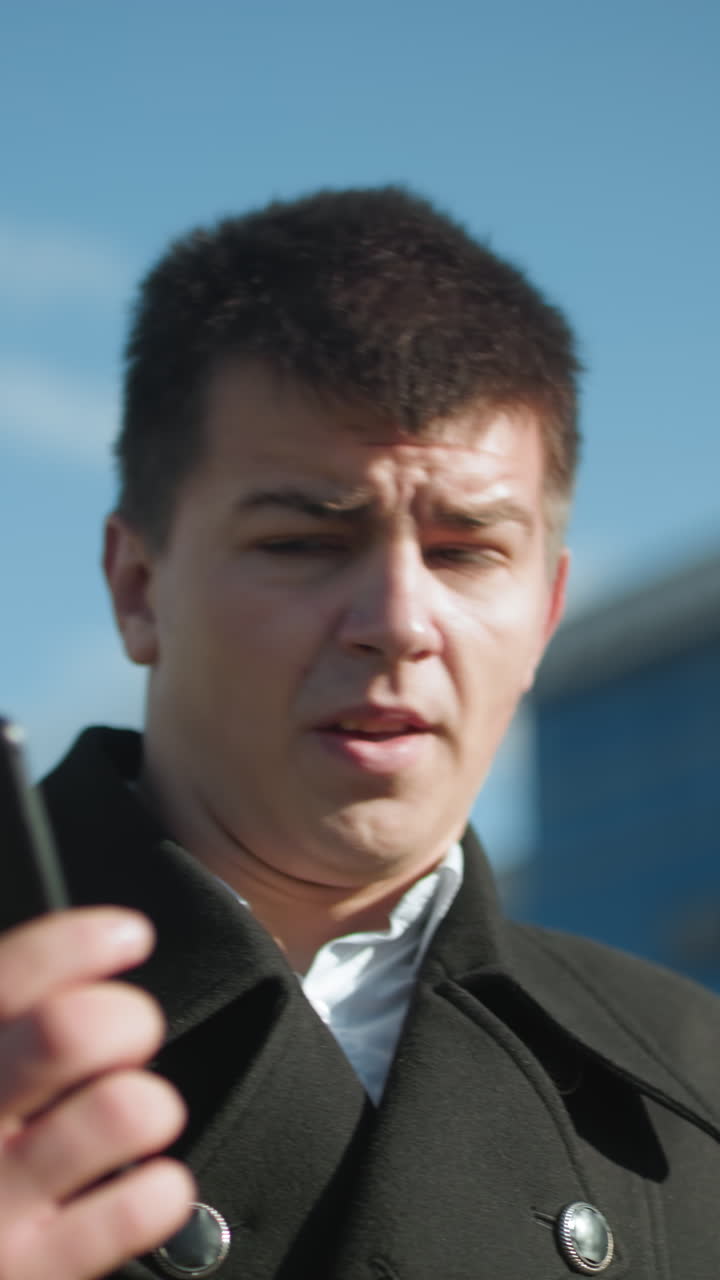 contratista con abrigo negro con botones metálicos participa en una llamada telefónica al aire libre, parece contemplativo, toma una respiración profunda bajo un cielo brillante con edificios modernos en un fondo borroso