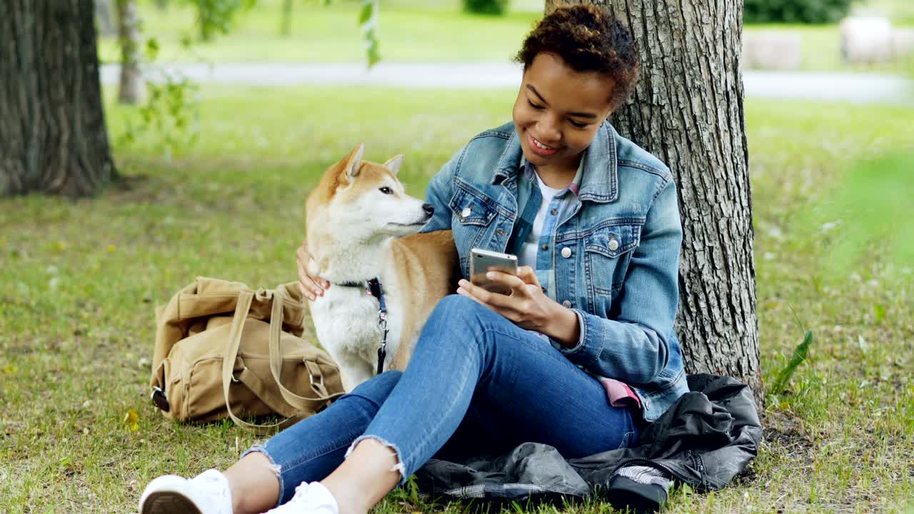 Sociable girl is using smartphone texting friends and stroking her adorable puppy while resting in park at weekend. Modern technology, people and animals concept.