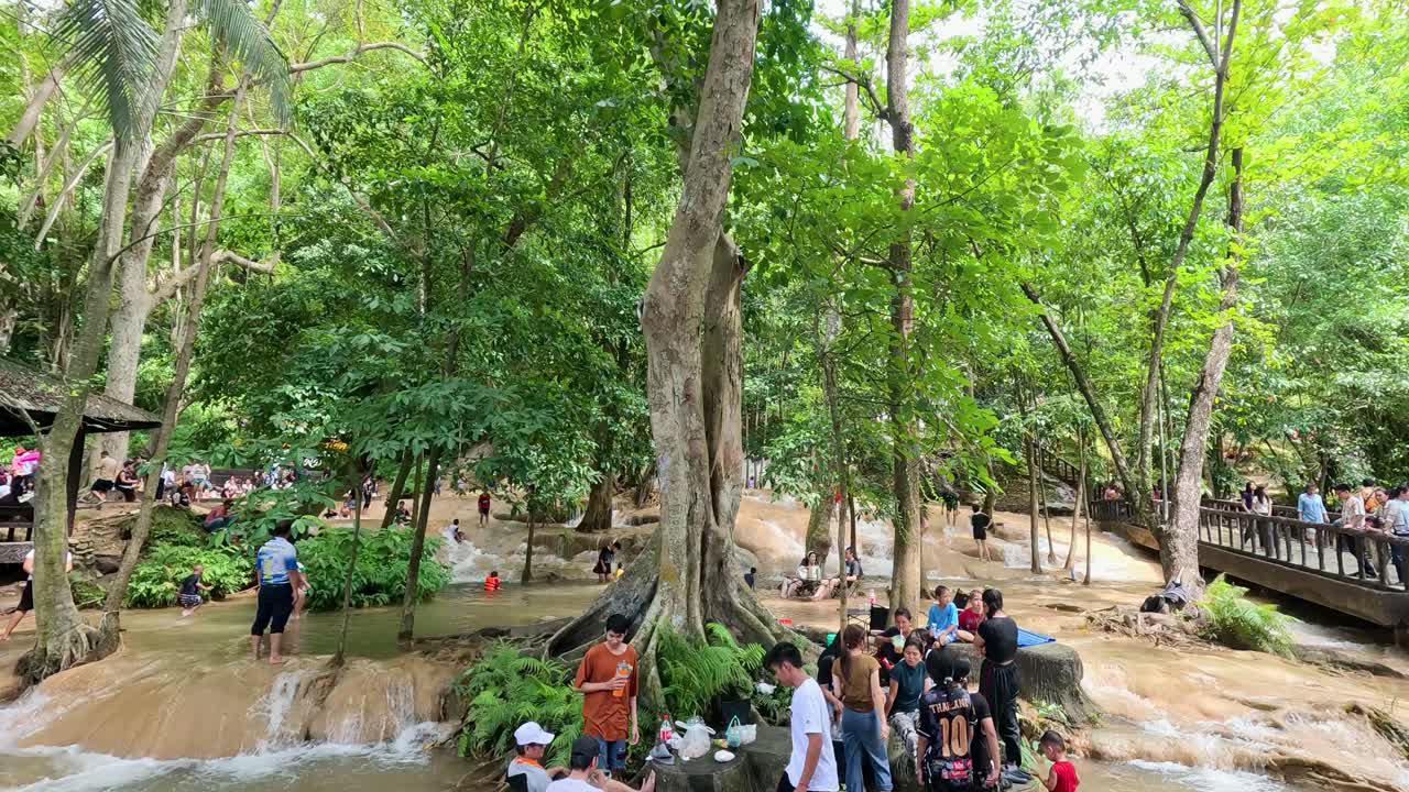 A group of people relaxes and interacts beside a small waterfall in a vibrant forest setting, with natural daylight and smooth camera movement
