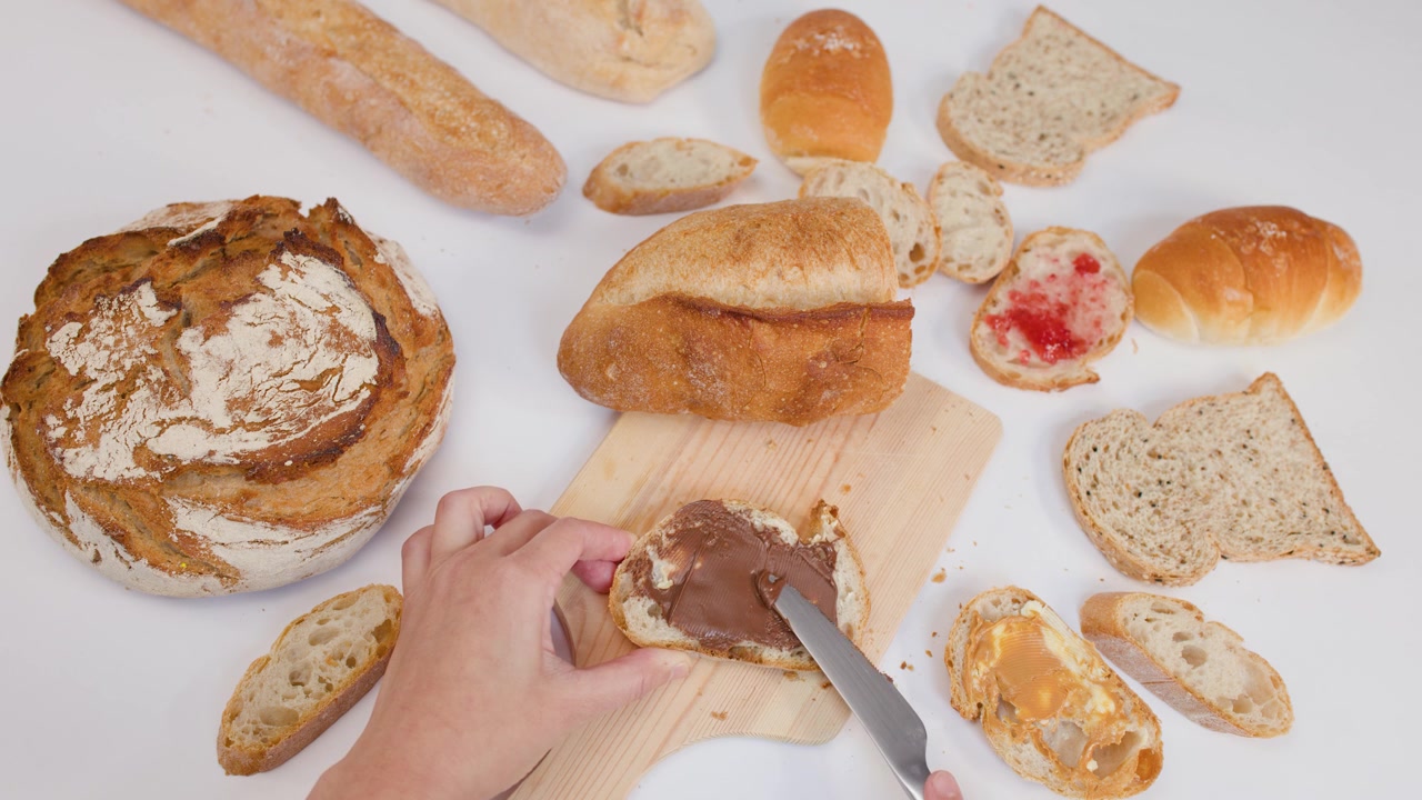 Ripresa dall'alto di mani che spalmano crema al cioccolato e nocciole sul pane, tra vari tipi di pane fresco.
