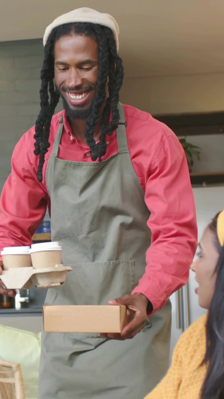 Video verticale: Cameriere con grembiule e berretto che porta l'ordinazione, posizionando una scatola da asporto e delle tazze sul tavolo in un caffè.