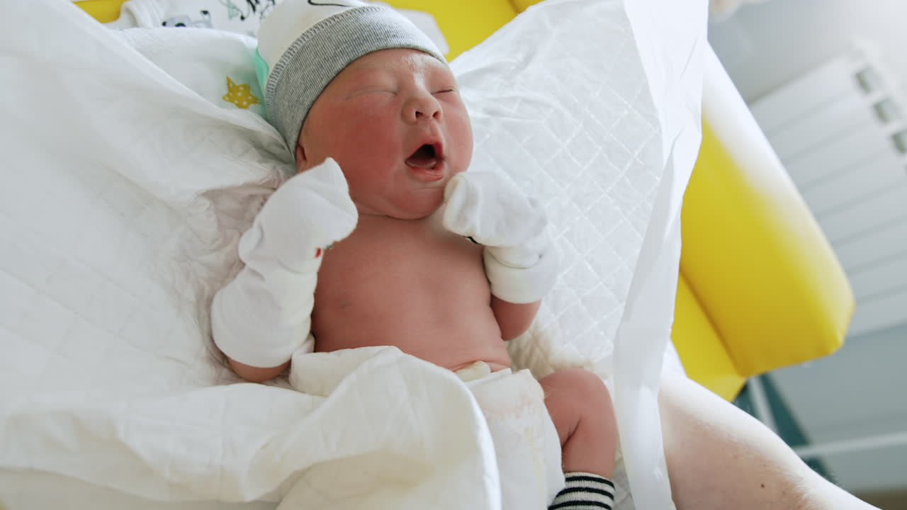 Tiny newborn baby in hat and mittens. Child in a wrap is taken into hands. Close up.