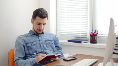 Young-businessman-sitting-by-the-computer-in-stylish-modern-office-and-taking-notes-using-his-pencil-and-notebook.-Computer,-phone-and-cup-on-the-table.-Shot-in-4k