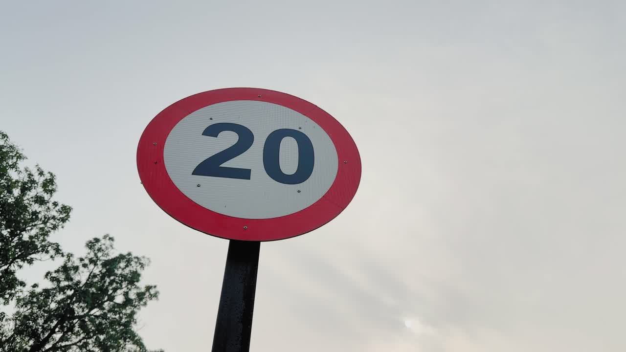 Cámara orbitando alrededor de una señal de tráfico circular de límite de velocidad de 20 montada sobre un poste; borde rojo y números destacados visibles sobre un cielo pálido y árboles a la luz del día