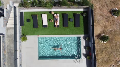 drone flying over an outdoor pool in bird's eye view - woman in swimming costume lying on sunbed and man swimming in pool - villa in greece crete with palm trees