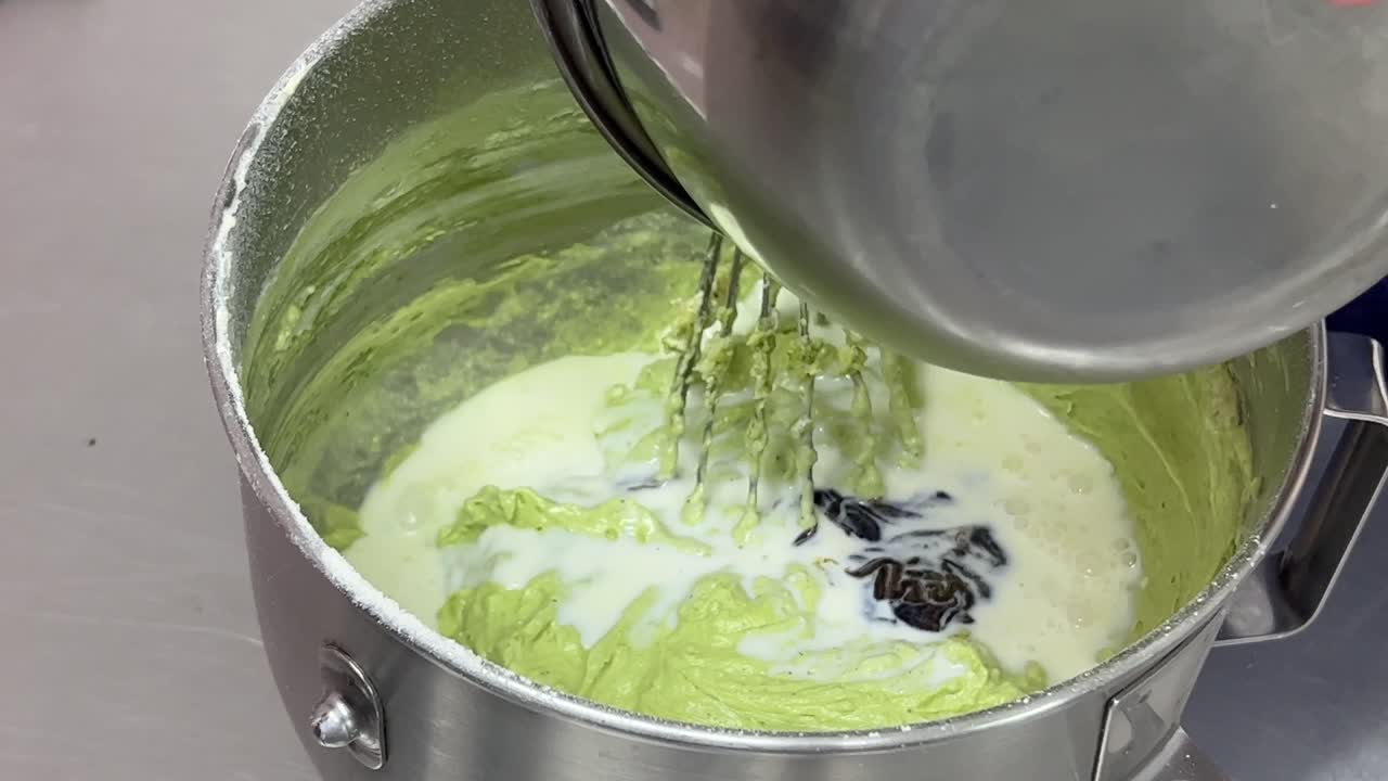 Pastry chef hand mixing with a whisk, patiently mixing matcha flavor cake mixture, slowly adding milk and vanilla essence into the bowl, close up shot