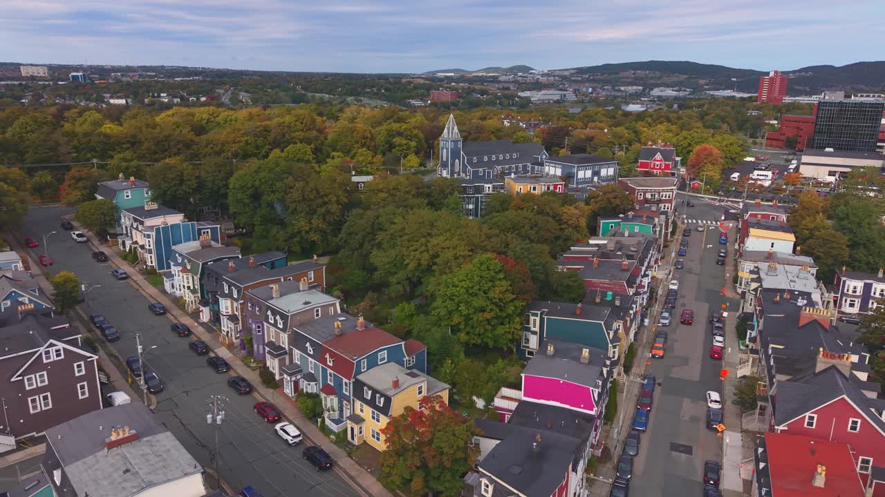 A high aerial shot over St. John's Newfoundland in autumn showcases colourful hillside homes, winding streets and a harbour bordered by lush forests under crisp clear skies