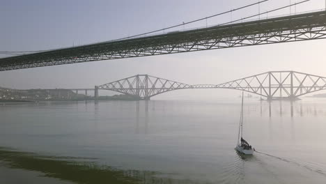 aerial footage of a sailing boat travelling beneath the old forth road bridge at south queensferry on a sunny day in west lothian, scotland