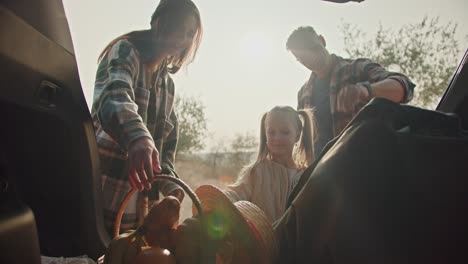 happy couple, a man with gray hair in a plaid shirt opens the trunk of the car and, together with his wife, takes out things for a picnic from there and, together with his little daughter, goes camping in the summer