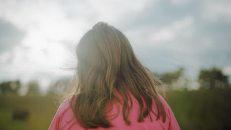 back view of woman in pink with long wavy hair cascading down, illuminated by warm sunlight, soft natural glow creates a serene countryside atmosphere with blurred trees and open sky