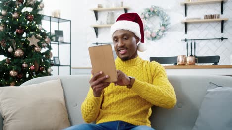 Portrait-Of-Joyful-Handsome-Young-Male-In-Good-Mood-Tapping-And-Typing-On-Tablet-While-Resting-In-Decorated-Room-Near-Glowing-Christmas-Tree-On-New-Year-Eve