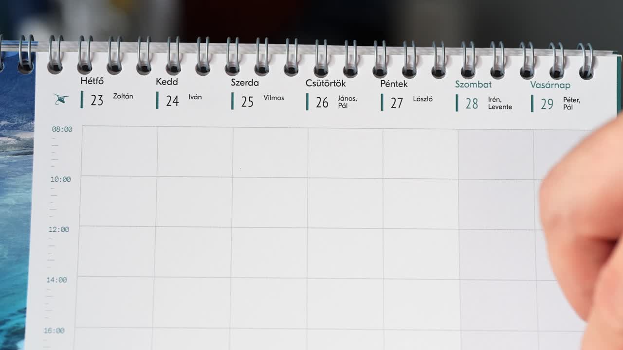 Close-up of a Hungarian weekly calendar showing the days from Monday to Saturday, with names associated with each date. A person is flipping the page, revealing part of the next week