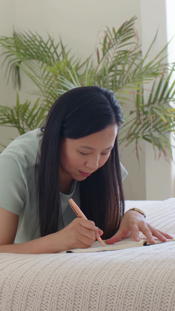 Video verticale: Donna con top verde chiaro a letto che scrive, usando una penna color oro rosa e un quaderno, mentre pianifica.
