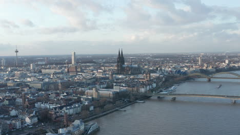 Backwards-fly-above-Rhine-river-in-city-at-dusk.-Busy-bridges-over-rippled-water-surface.-Gothic-Kolner-Dom-towering-above-surrounding-development.-Cologne,-Germany