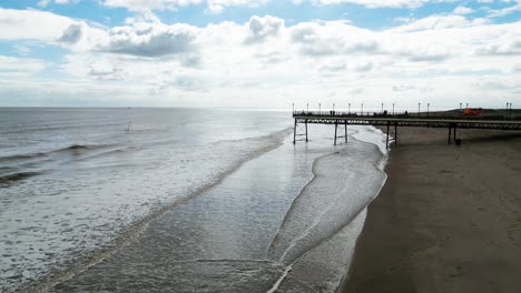 Typical-English-seaside-resort,-shot-using-a-drone-flying-into-the-sun-giving-a-aerial-viewpoint-with-a-wide-expanse-of-beach-with-a-pier-and-crashing-waves-1