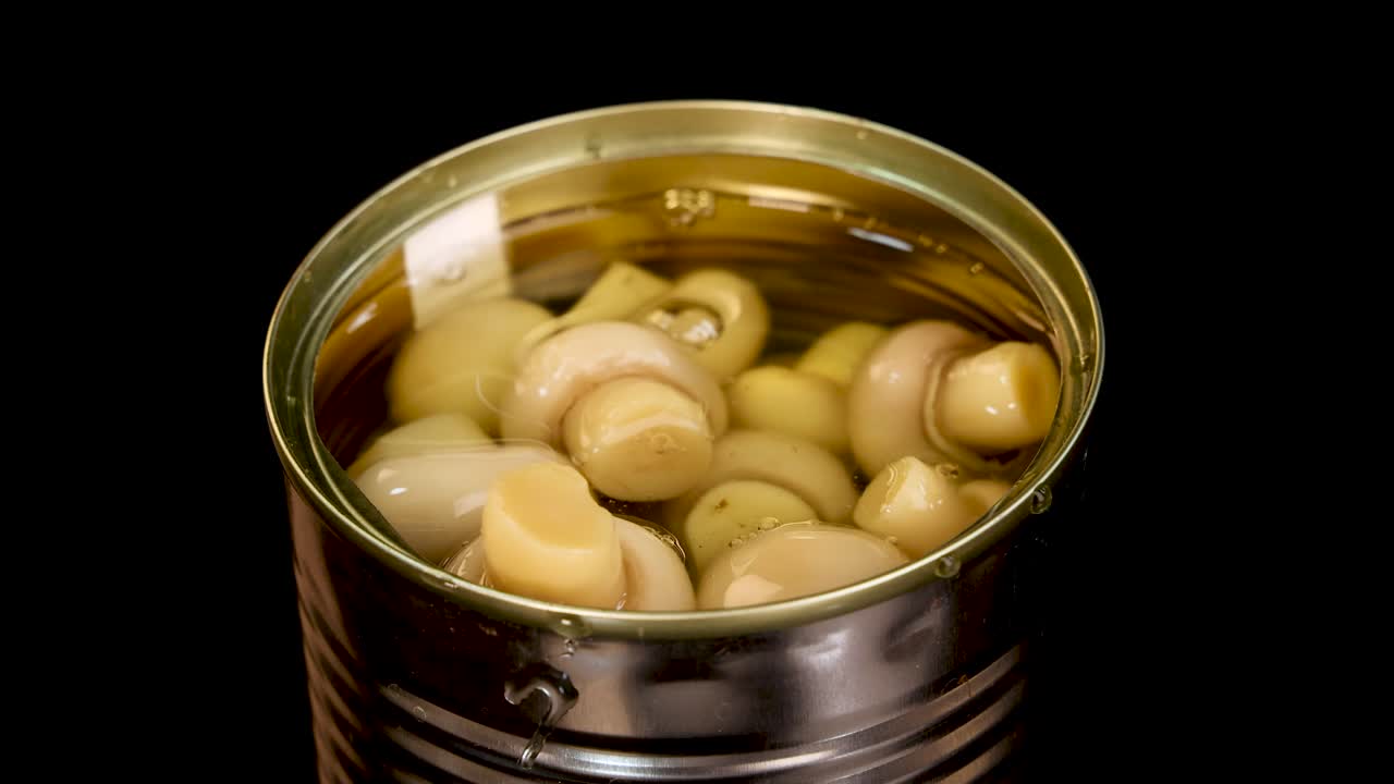 Whole champignon mushrooms fall into an open can filled with liquid, creating a splash. Studio lighting, black background, and static overhead camera enhance clarity