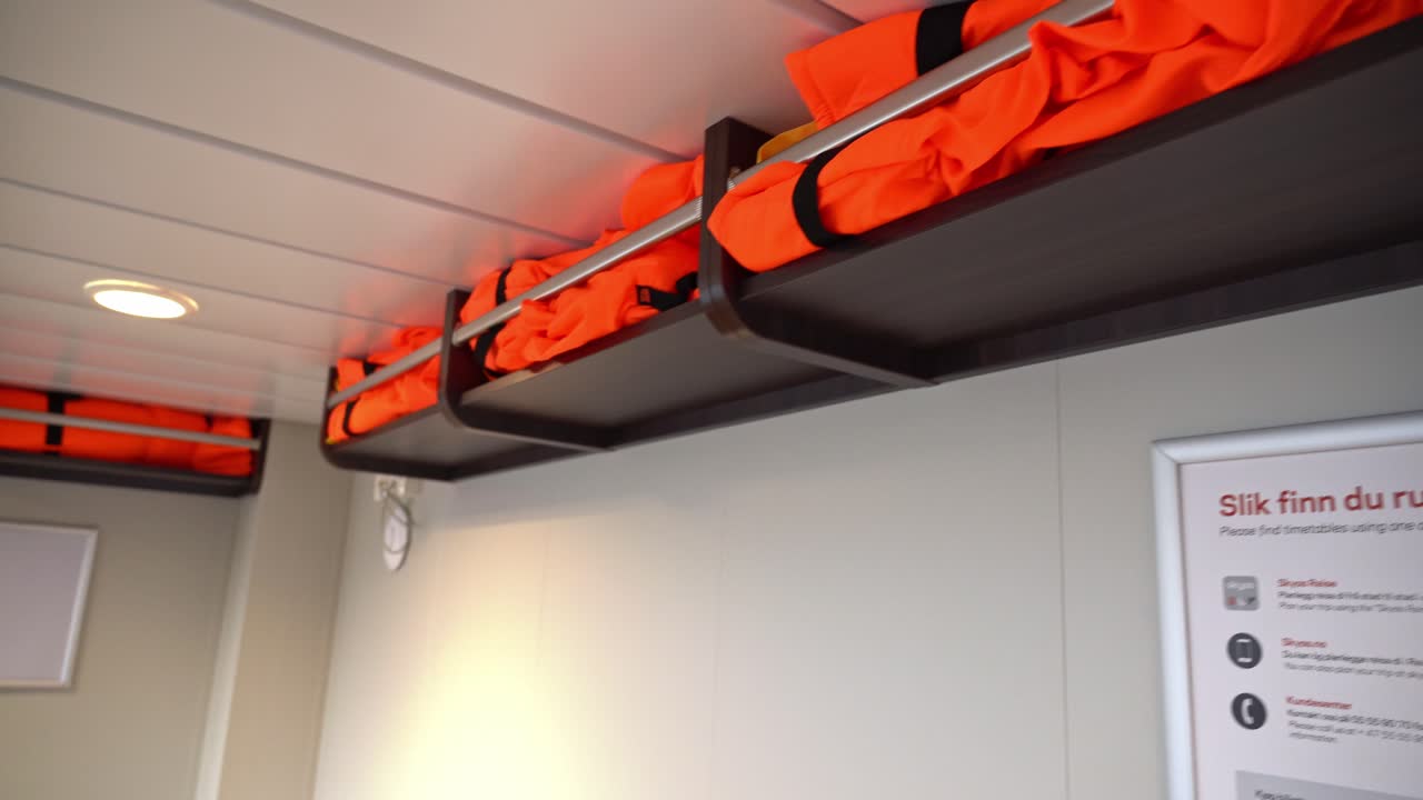 Lifejackets stored in shelves over seats onboard passenger ship - Camera looking straight at closest shelf before panning left and revealing passenger area