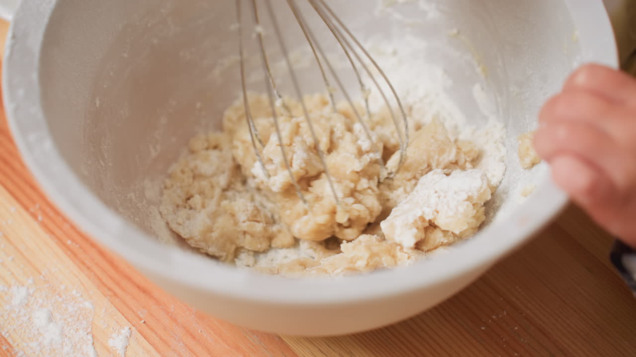 Close up of child mixing flour into dough with whisk, struggling to stir thick mixture inside bowl, hands visible holding whisk tightly while attempting to blend ingredients